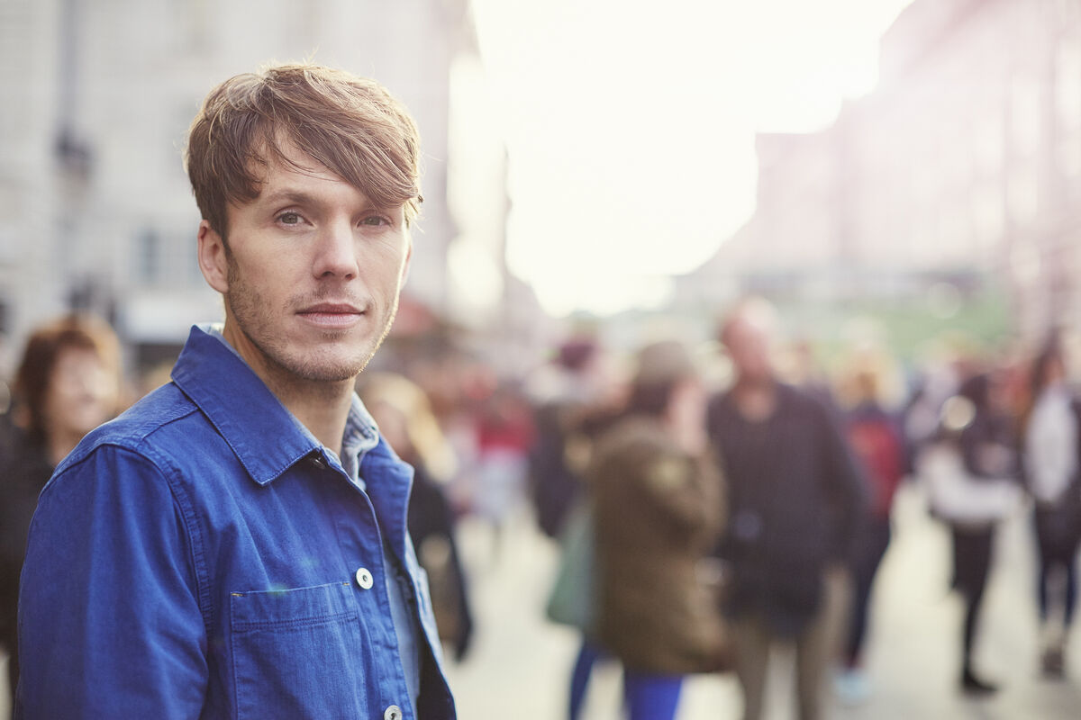 A man with an introspective expression standing in an urban street, soft afternoon light.