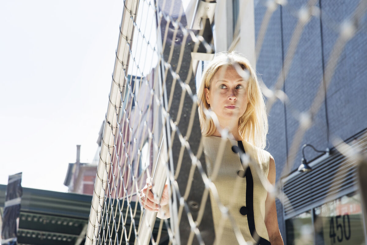 A woman standing thoughtfully outside, framed by a metal fence, in soft morning light.