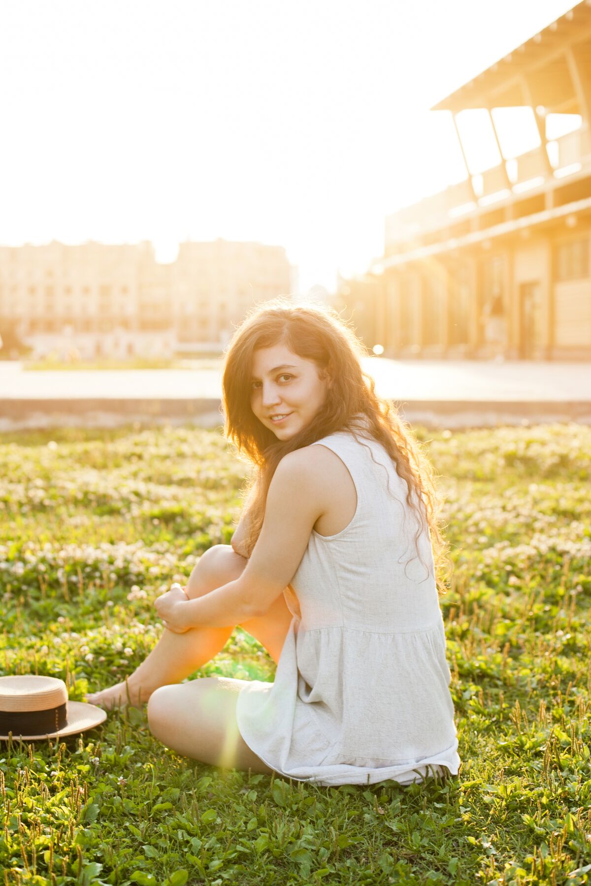 A woman in a sundress and hat sitting reflectively on grass, soft summer light.